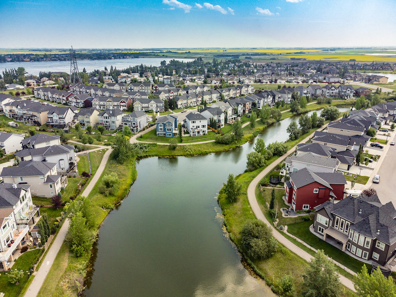 Drone Shot of Suburban Neighborhood and Waterway in Chestermere An elevated aerial view showcasing a dense residential subdivision in Chestermere, Alberta. Modern suburban houses line a winding green waterway with walking paths and green space under a bright sky with light clouds.