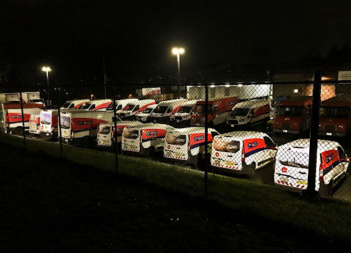 A night shot of a large fleet of commercial delivery vans parked side-by-side in a chain-link fenced lot. Bright overhead floodlights illuminate the branded red, white, and blue vehicles parked outside a depot facility.