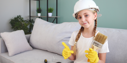 A young girl wearing a white hardhat, safety gloves, and holding a paintbrush on a living room sofa, symbolizing DIY home renovation and painting trends in Canada.